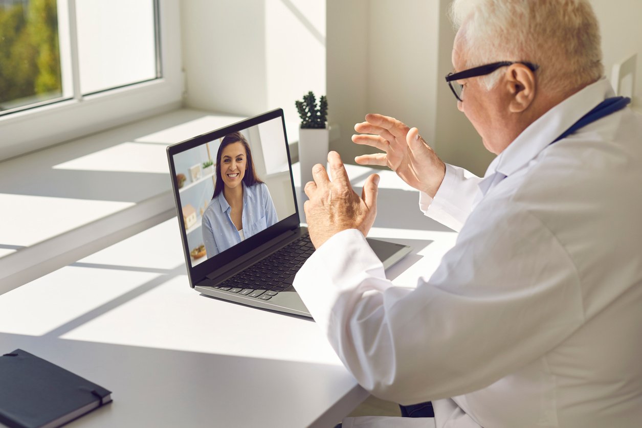 Senior Doctor Sitting at Desk in Office and Giving Online Consultation to Happy Woman